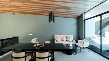 A modern living room with a black dining table, beige chairs, a sofa with cushions, and a large window offering a view of a pool and outdoor area.