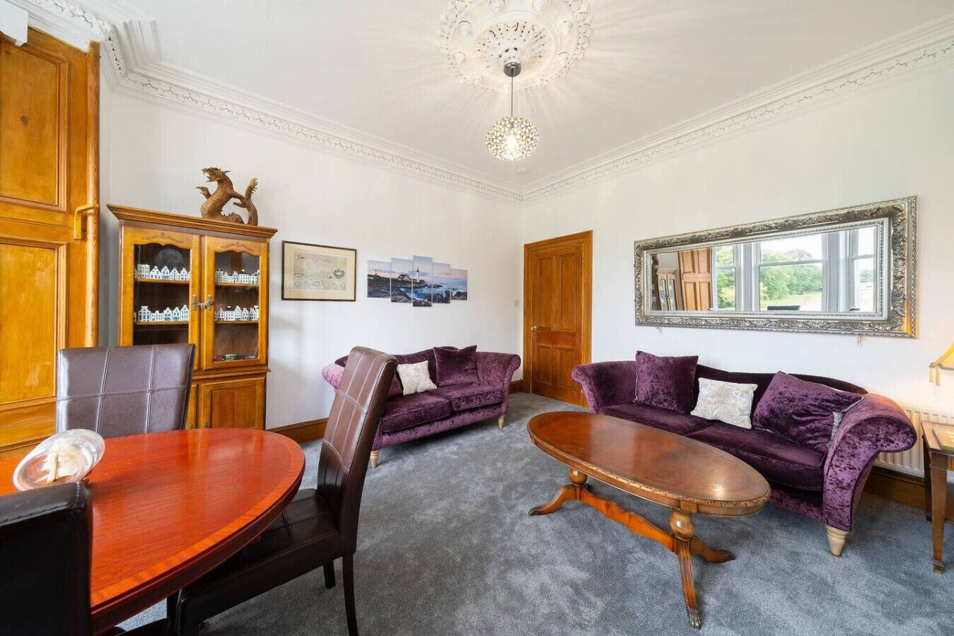 A room with a wooden dining table, two purple sofas, a wooden cabinet with glass doors, and a large mirror on the wall.