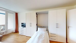 Une chambre à coucher avec un lit, un téléviseur posé sur une commode, une armoire et une fenêtre avec des rideaux.