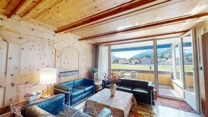 Een ruime woonkamer met houten balken in het plafond, grote ramen met uitzicht op een bergachtig landschap, en diverse zithoeken, waaronder een blauwe bank, een zwarte bank en een koffietafel met een patroon.