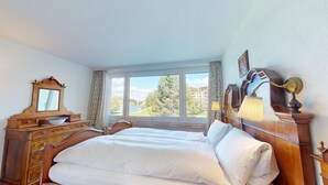 Een slaapkamer met een groot bed, een houten dressoir en een raam met uitzicht op groen en gebouwen.