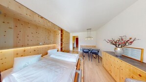 A modern hotel room with two beds, a wooden headboard, a dining area with a table and chairs, and a wooden cabinet with a vase of flowers. - Well-kept holiday apartament house in a prime location in St. (St. Moritz)