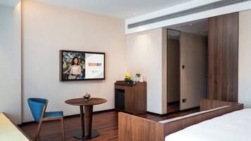 A hotel room with a bed, a desk with a vase of flowers, a chair, a small round table, and a framed picture on the wall.