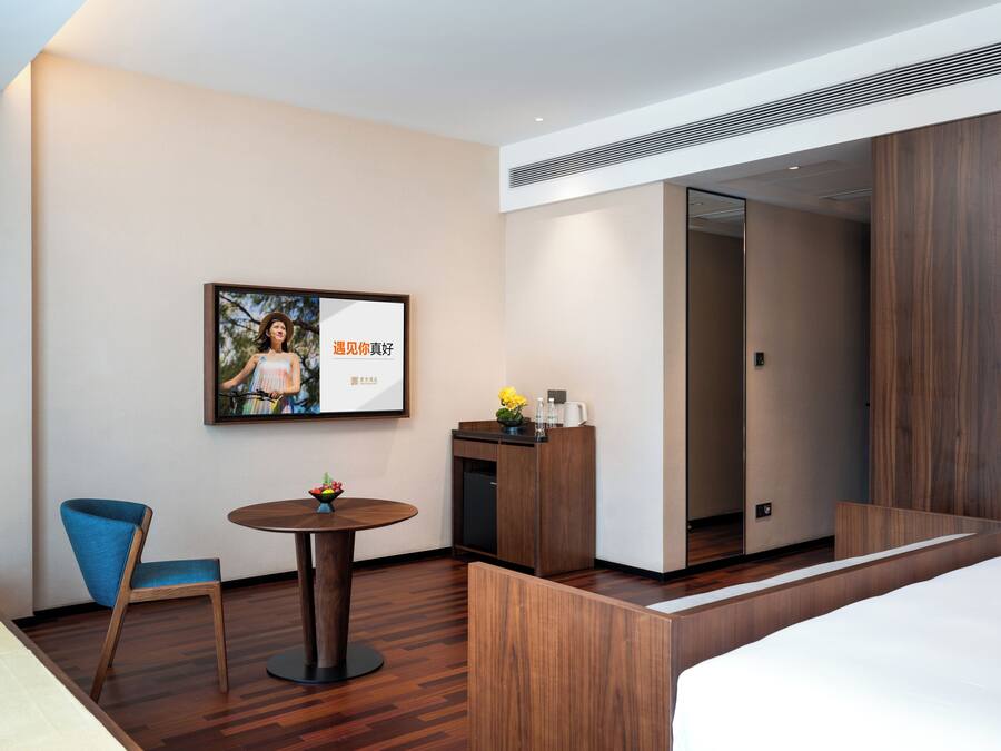 A hotel room with a bed, a desk with a vase of flowers, a chair, a small round table, and a framed picture on the wall.