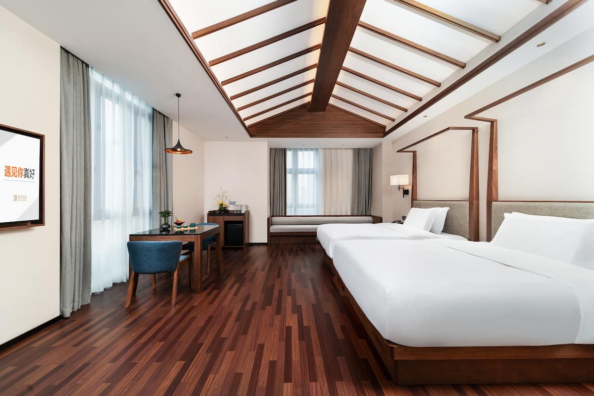 A hotel room with a wooden floor, a bed, a desk with a chair, a window with curtains, and a ceiling with wooden beams.