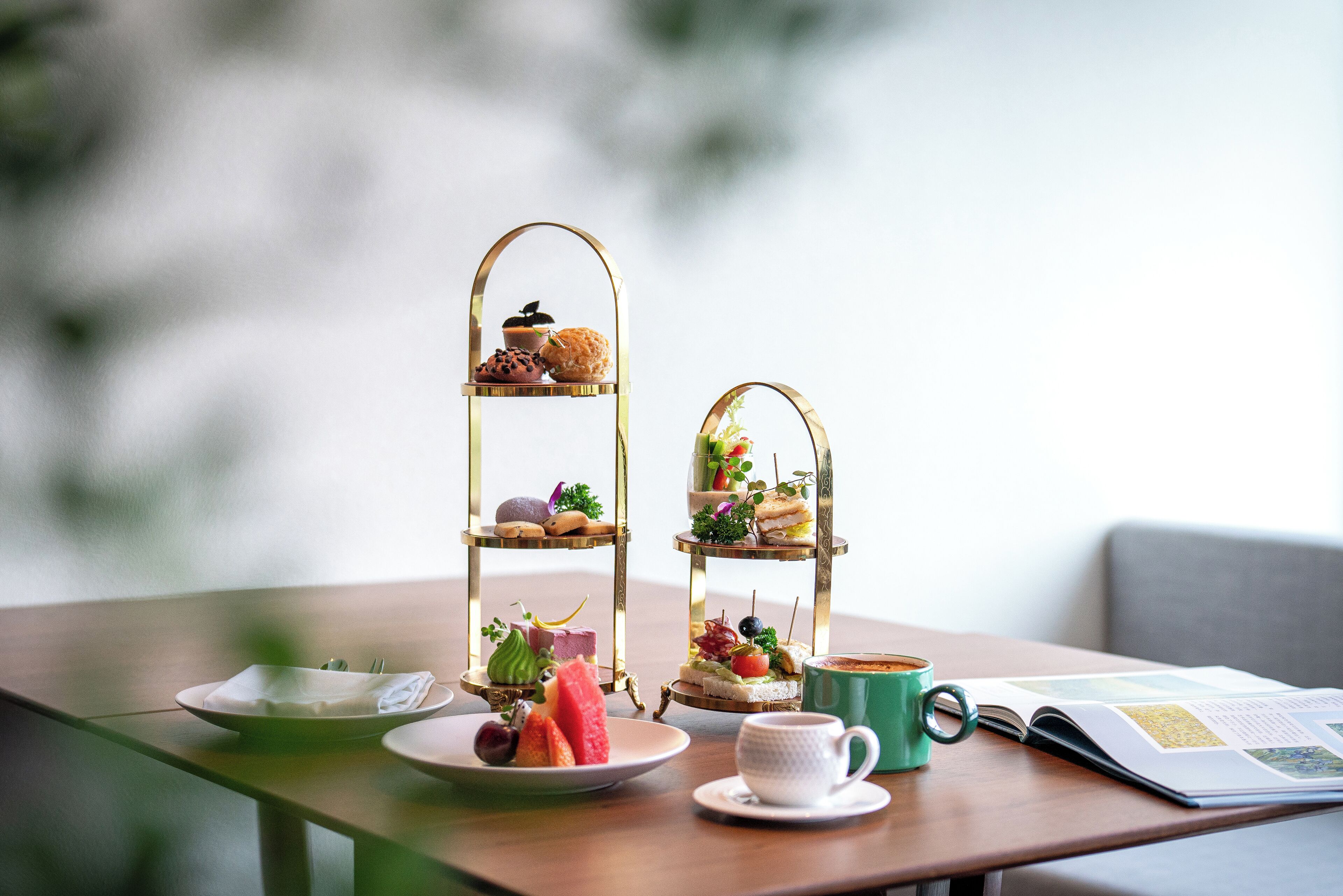 A table with a tiered dessert stand, a plate with a napkin, a cup of coffee, and a magazine.