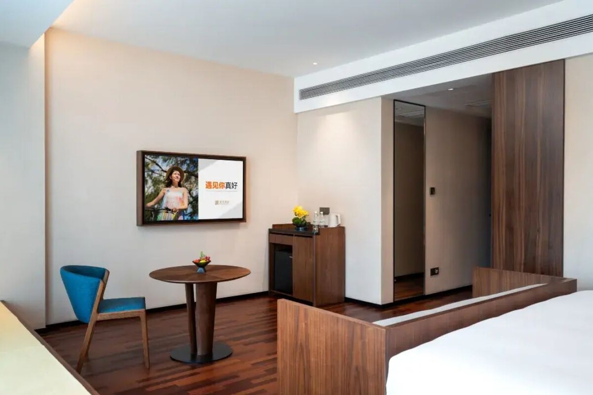 A hotel room with a bed, a desk, a chair, a small table with flowers, a framed picture on the wall, and a mirror.