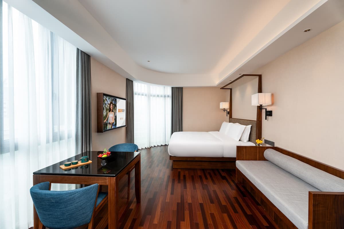 A hotel room with a bed, a sofa, a dining table with chairs, a TV, and a large window with curtains.