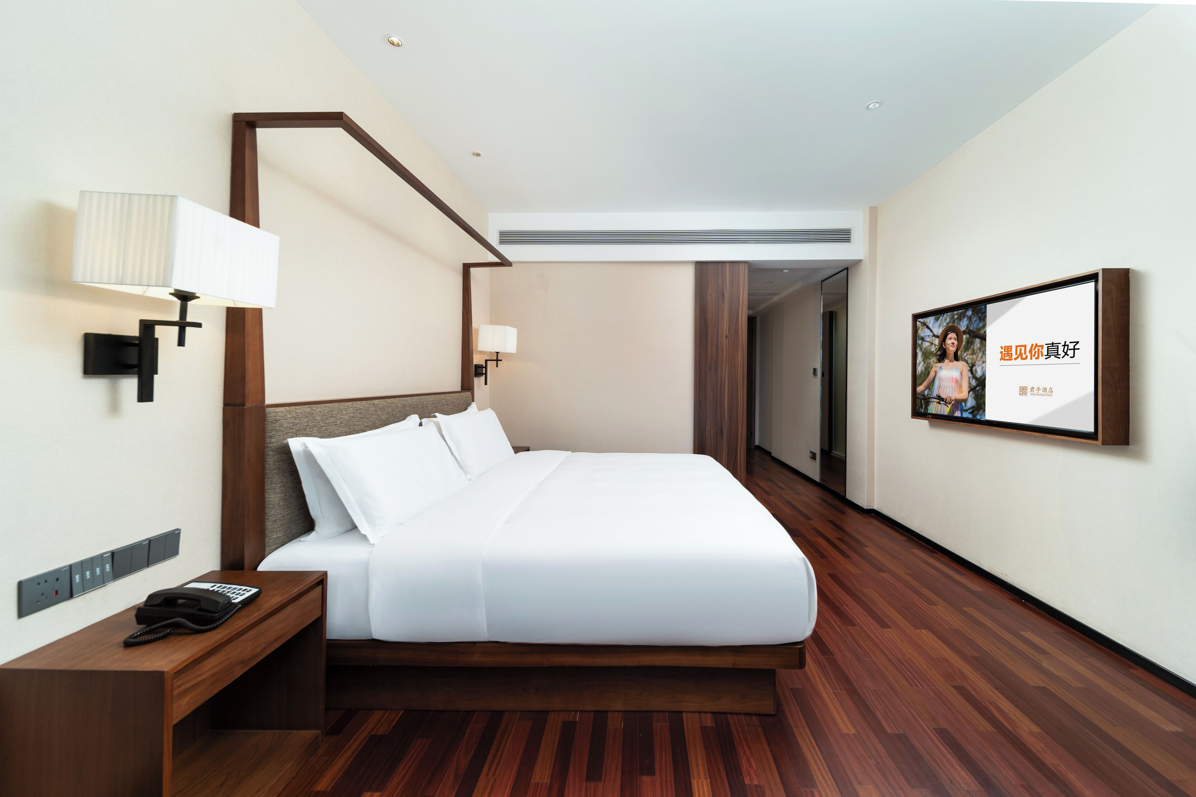 A modern hotel room with a wooden floor, a large bed with white linens, a bedside table with a phone, and a wall-mounted TV displaying a photo.