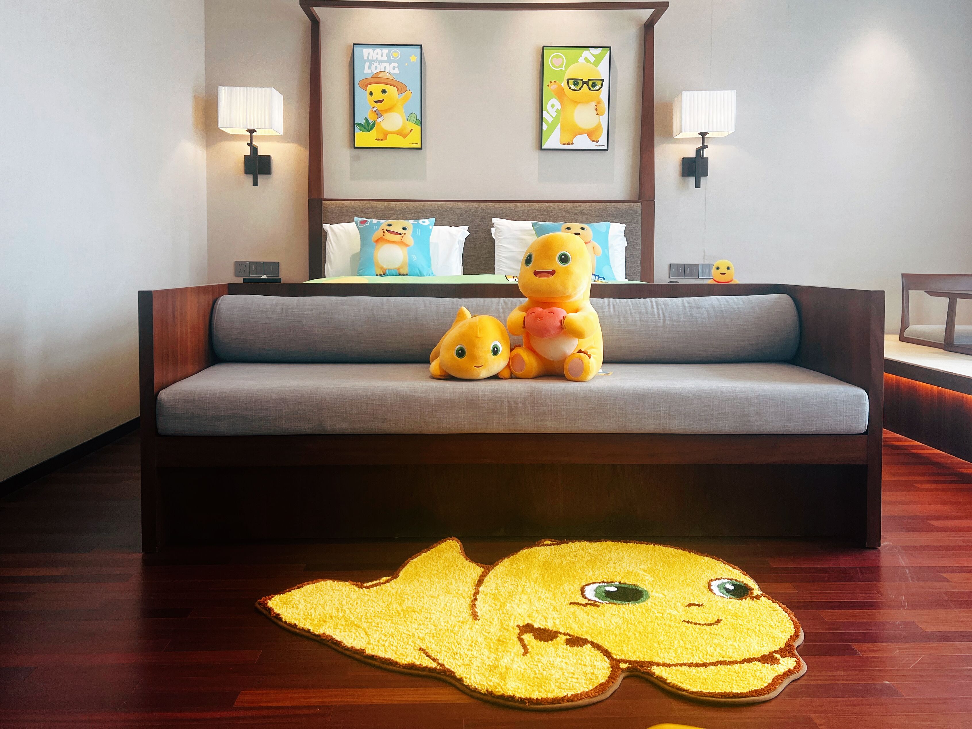 Quarto de hotel com um sofá cinzento, dois brinquedos macios em cima e um tapete decorativo grande no chão.