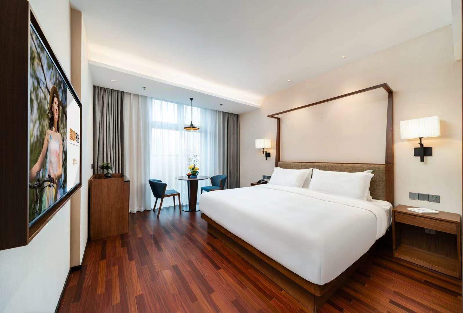 Habitación de hotel con una cama grande, un escritorio con una silla, una mesita con un florero y una ventana con cortinas.