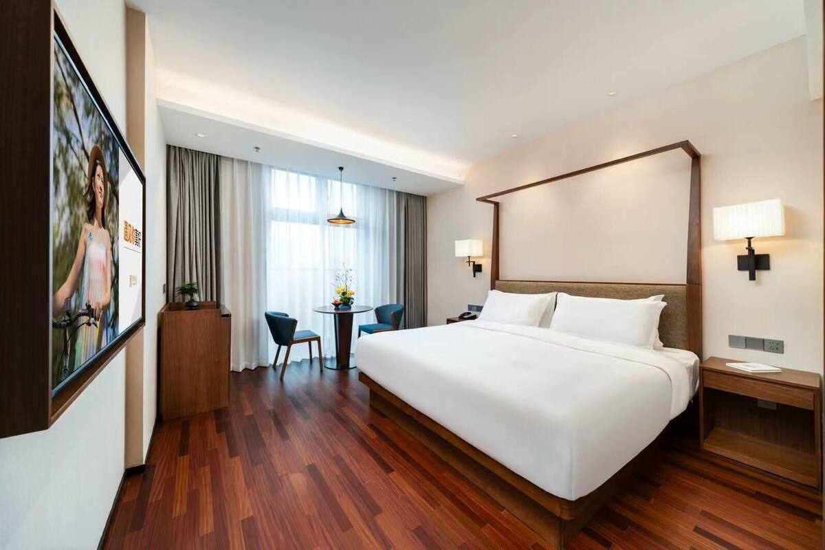 A hotel room with a large bed, a desk with a chair, a small table with a vase, and a window with curtains.