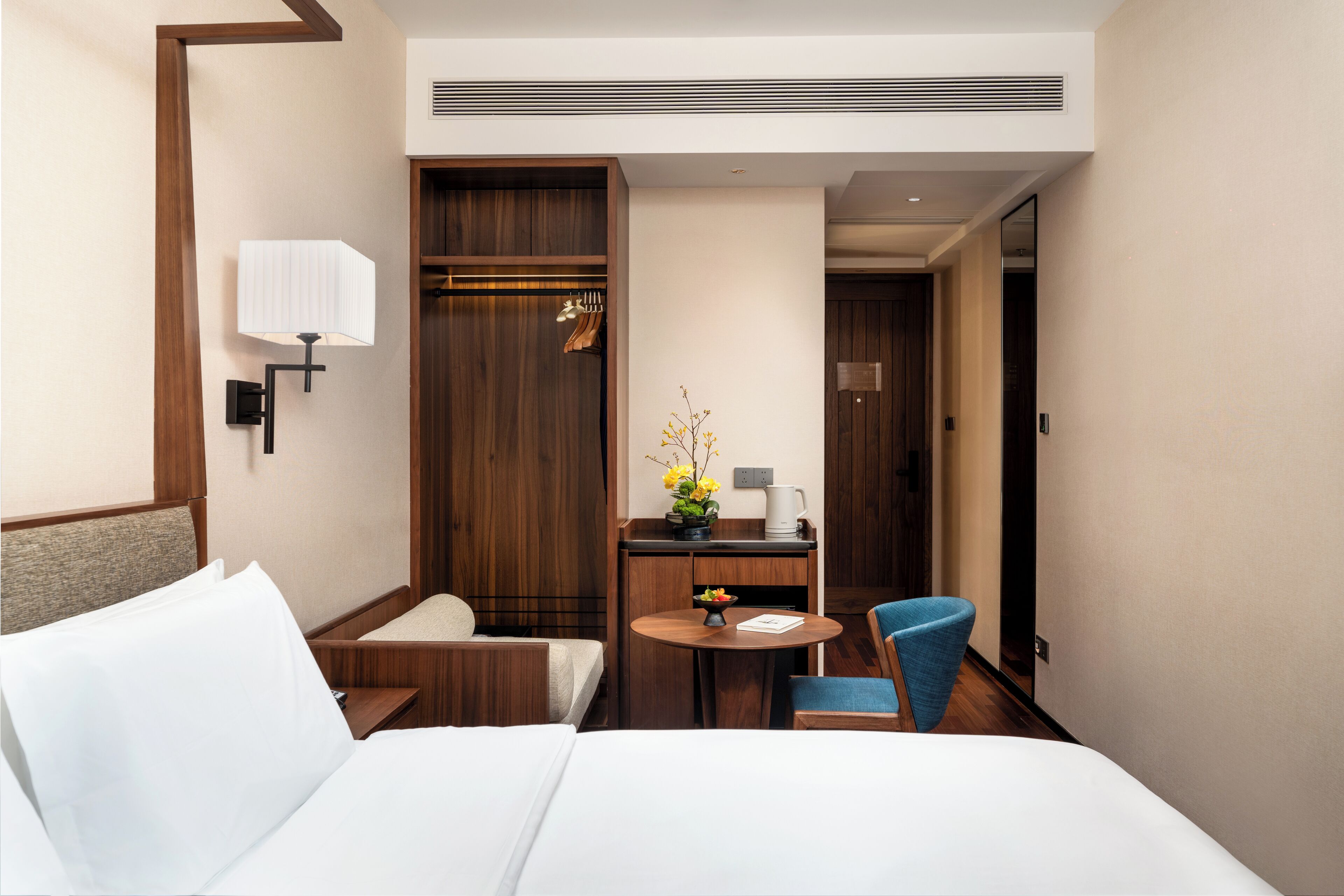 A hotel room with a bed, a desk with a chair, a small table with a vase of flowers, and a coat rack.