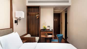 A hotel room with a bed, a desk with a chair, a small table with a vase of flowers, and a coat rack.
