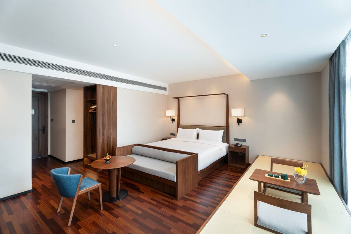 A modern hotel room with a large bed, a small table, a chair, and a bedside table with lamps.