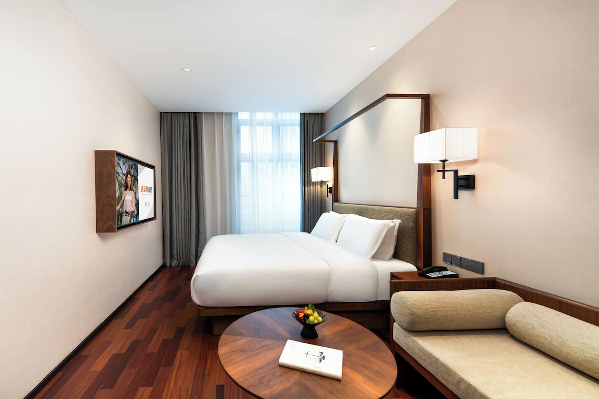 A hotel room with a large bed, a sofa, a round coffee table with a book and fruit, a bedside lamp, a phone, and a framed picture on the wall.