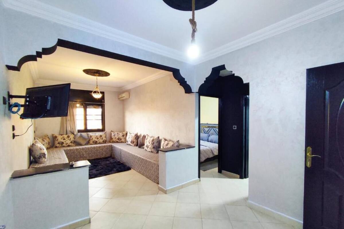 A living room with a sofa set, a flat-screen TV mounted on the wall, and a ceiling light fixture.
