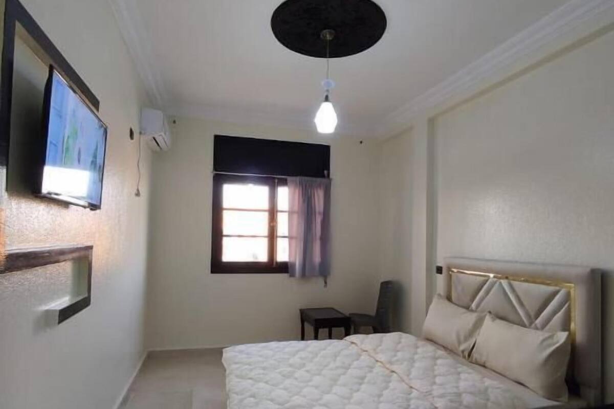 A bedroom with a bed, a TV mounted on the wall, a ceiling light, and a window with curtains.