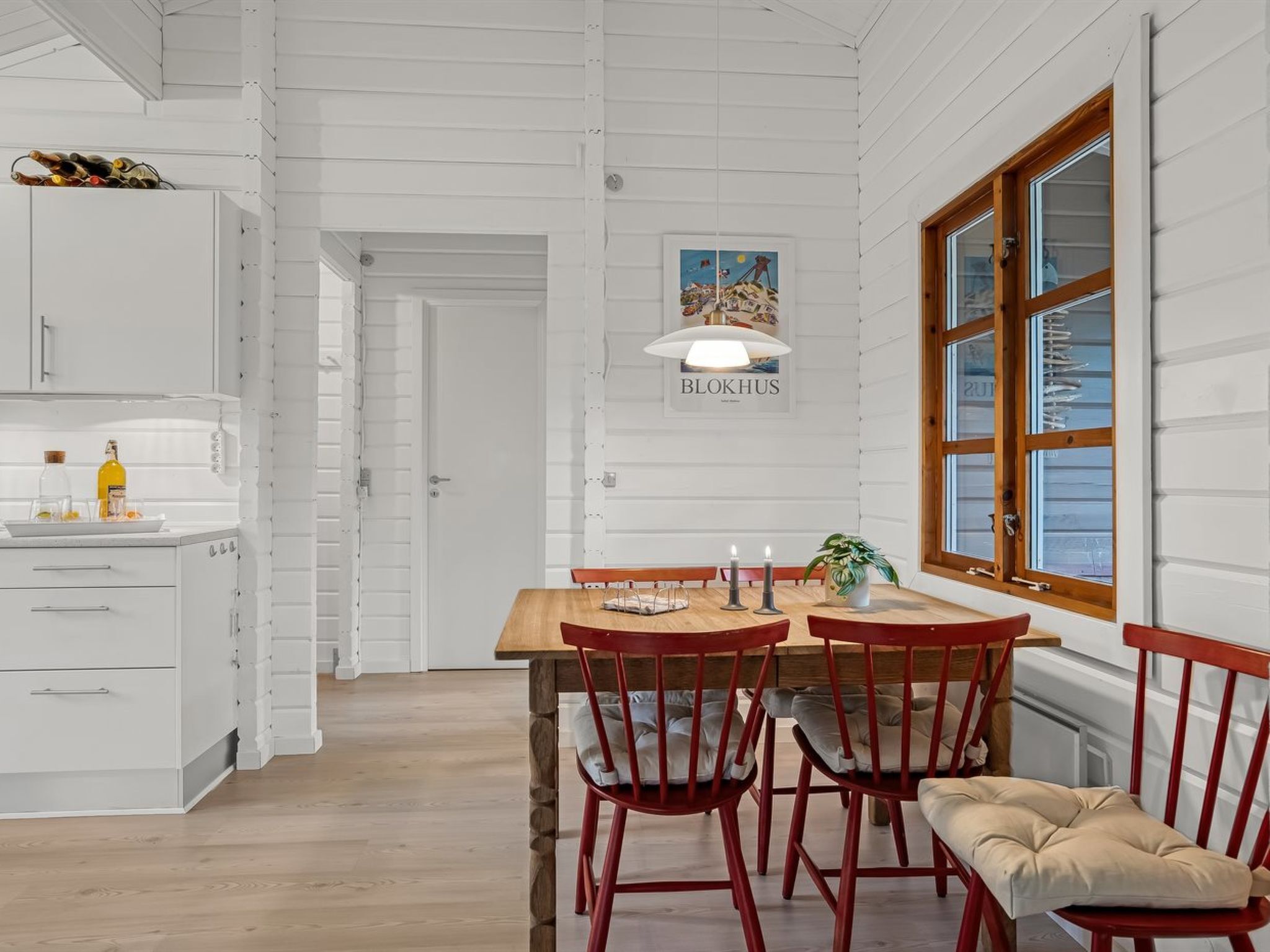 A dining area with a wooden table and red chairs, a white kitchen with cabinets, and a window with wooden frames.