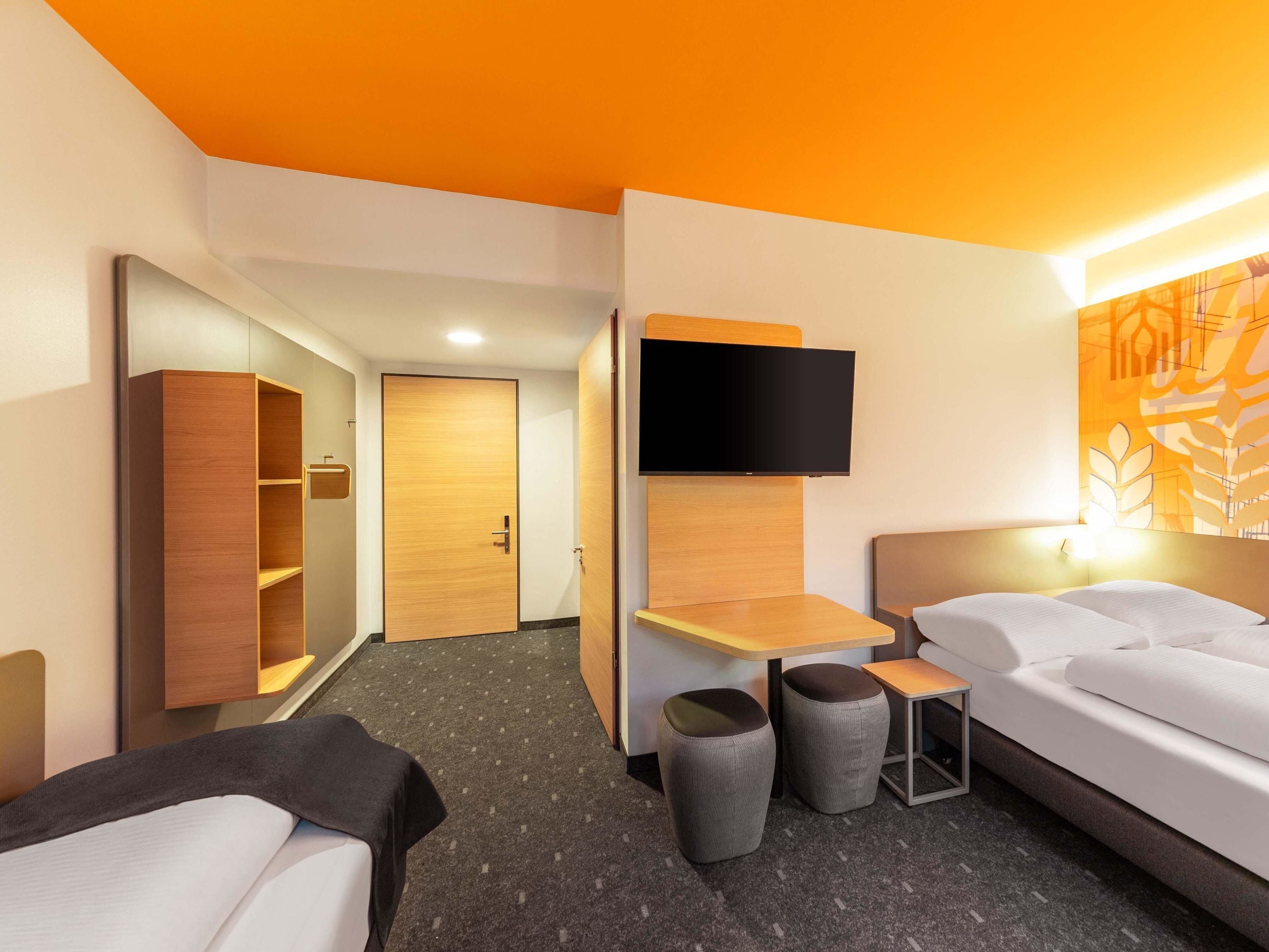 A modern hotel room with a bed, a desk, a wardrobe, and a TV mounted on the wall.