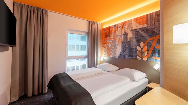 Una habitación de hotel con cama, escritorio, televisión y ventana con cortinas.