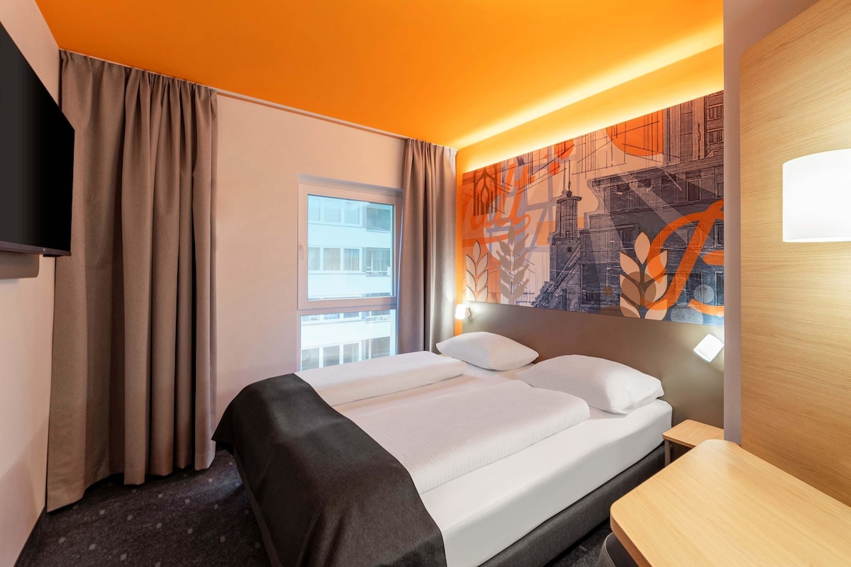 Une chambre d’hôtel avec un lit, un bureau, une télévision et une fenêtre avec des rideaux.