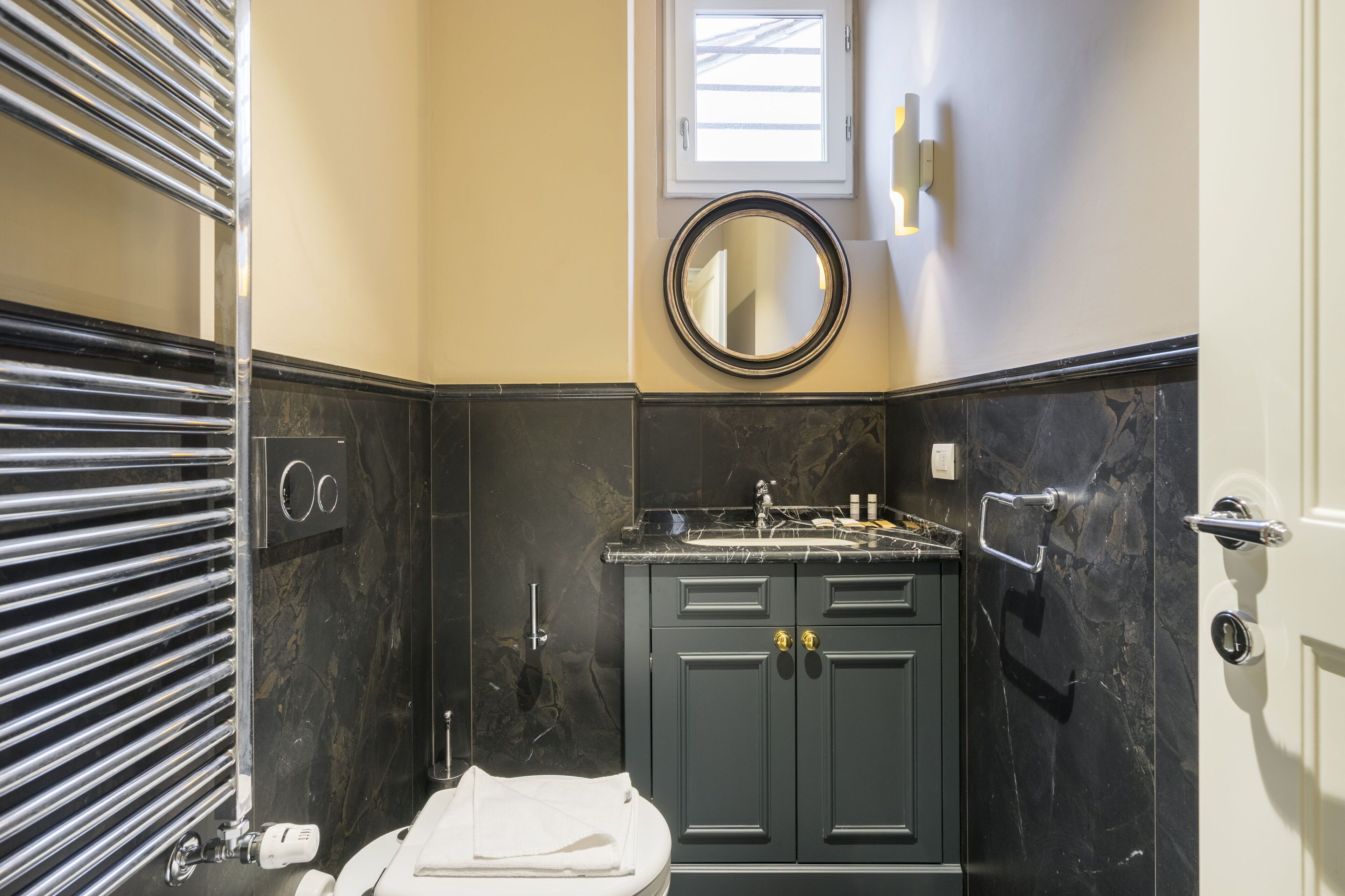 Une salle de bain avec un meuble-lavabo en marbre noir, un miroir rond, une serviette sur le sol et une fenêtre au-dessus du miroir.