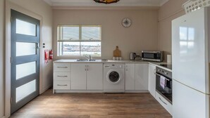 A modern kitchen with a washing machine, microwave, oven, and a large window with blinds. - Cosy and Comfortable Unit with Parking & Wifi (Devonport)