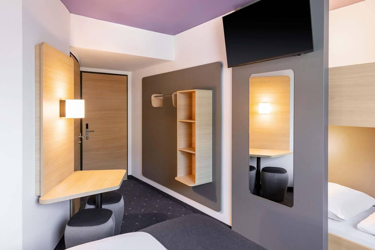 A modern hotel room with a bed, a desk, a chair, a TV, and a shelf with storage.