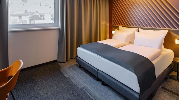 Een moderne hotelkamer met een groot bed, uitzicht op gebouwen door het raam en een bijzonder plafondontwerp.