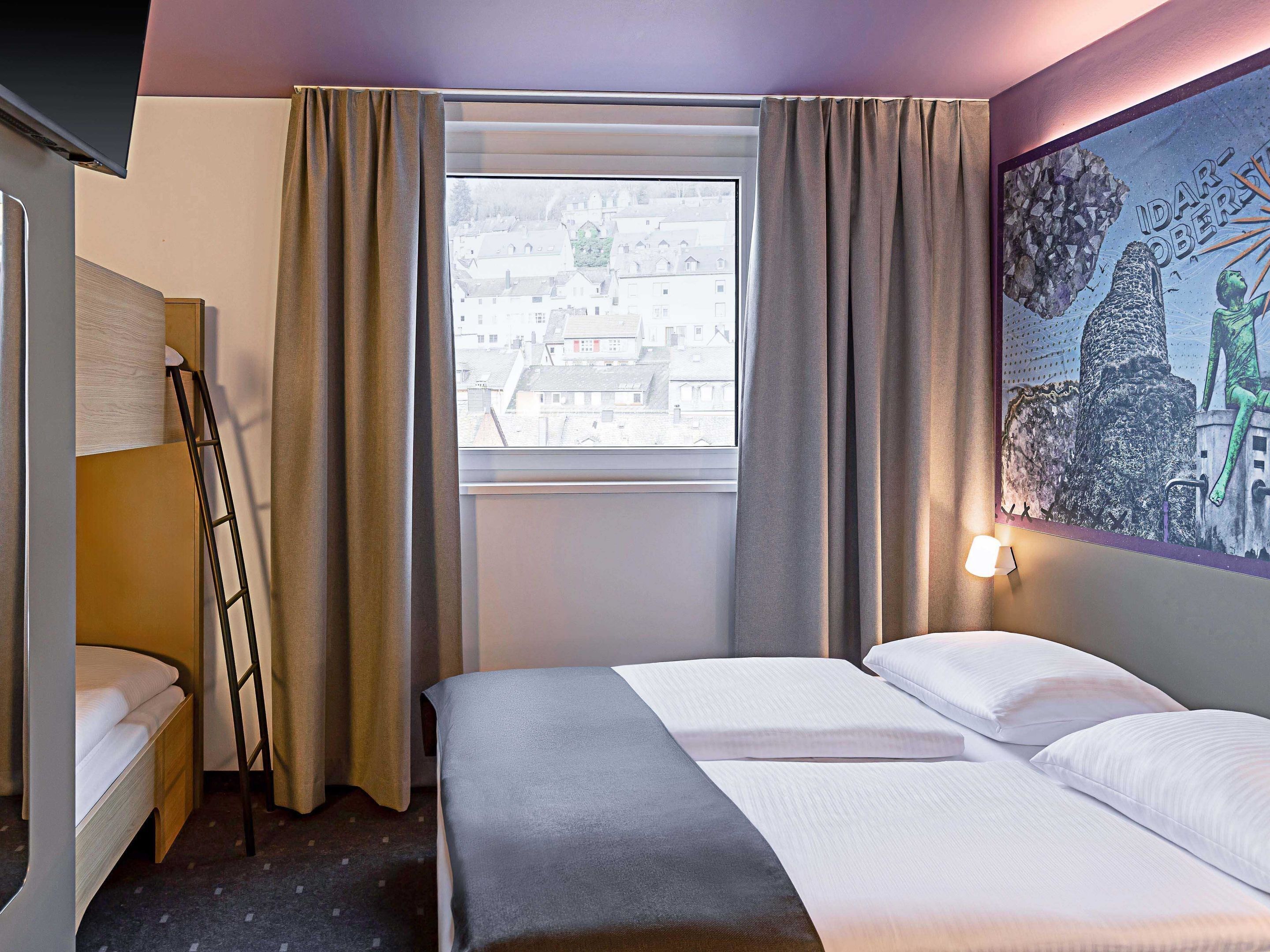 A hotel room with a bed, a desk, a chair, a ladder, and a window with curtains.