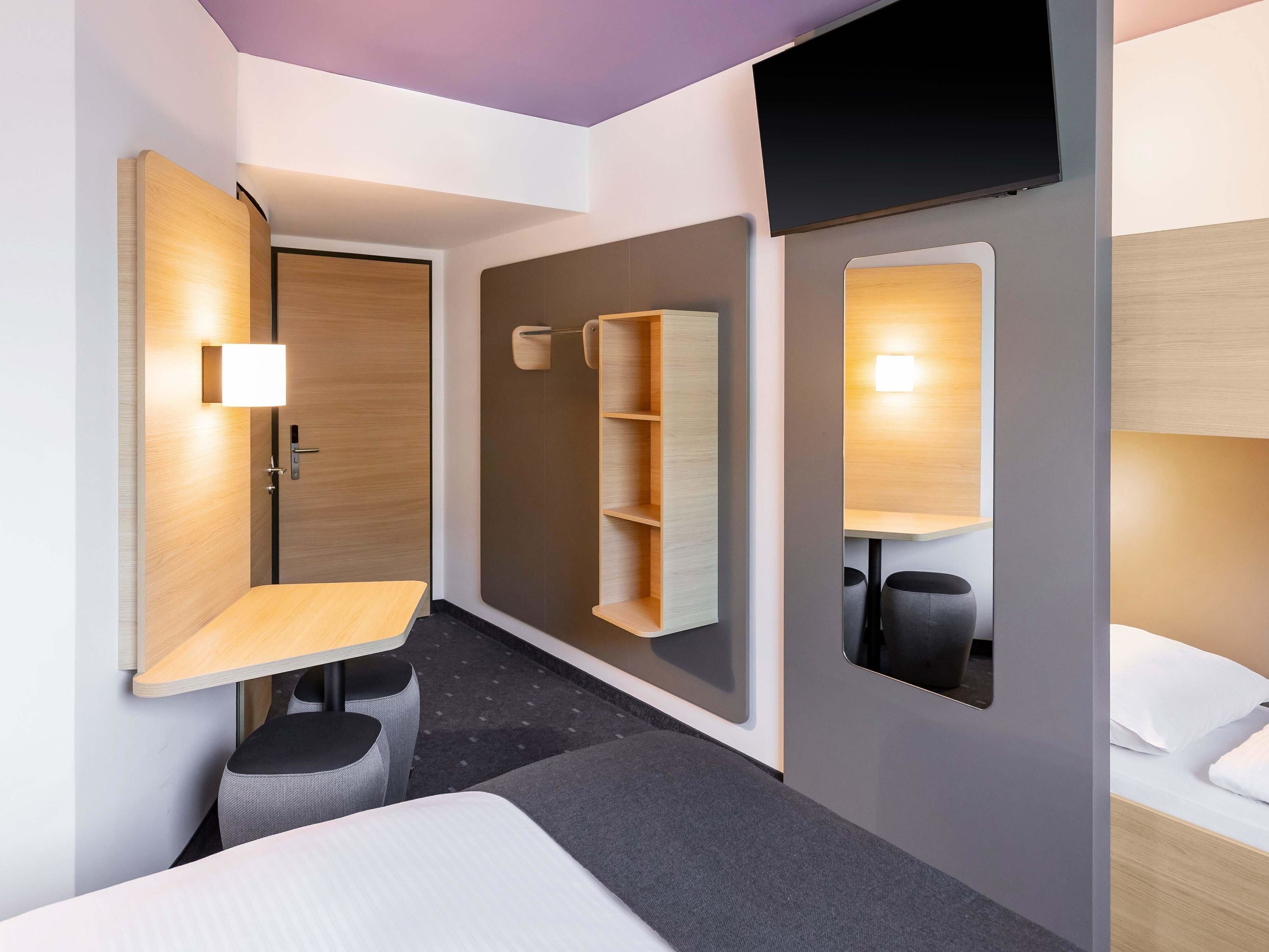 Ein modernes Hotelzimmer mit Bett, Schreibtisch, Sessel, Fernseher und einem Regal mit Stauraum.