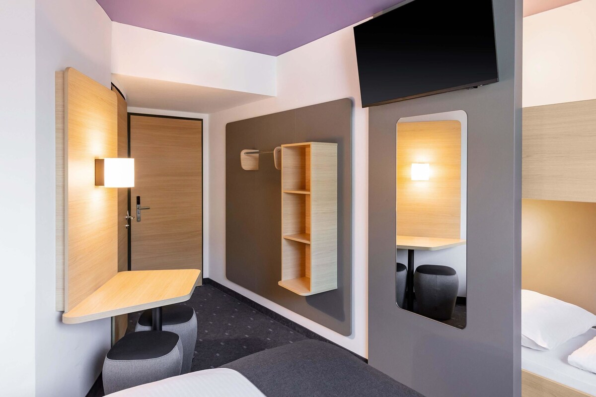 Moderni hotellihuone, jossa on sänky, työpöytä, tuoli, televisio ja säilytystilaa tarjoava hylly.