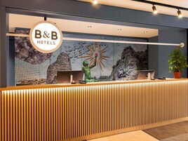 Una recepción con un cartel de B&B Hotels, un mural con un sol y montañas, y una planta en maceta.