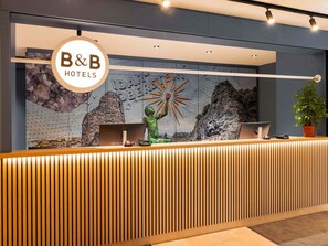 Una recepción con un cartel de B&B Hotels, un mural con un sol y montañas, y una planta en maceta.