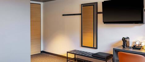 Une chambre d’hôtel moderne équipée d’un téléviseur à écran plat fixé au mur, d’un petit bureau avec une machine à café et d’un fauteuil confortable de couleur orange.