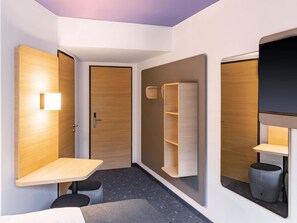 A modern hotel room with a wooden door, a small table, a built-in shelf, and a TV mounted on the wall. - B&B HOTEL Idar-Oberstein (Idar-Oberstein)