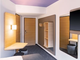 Habitación de hotel moderna con puerta de madera, una mesita pequeña, estante empotrado y televisor montado en la pared.