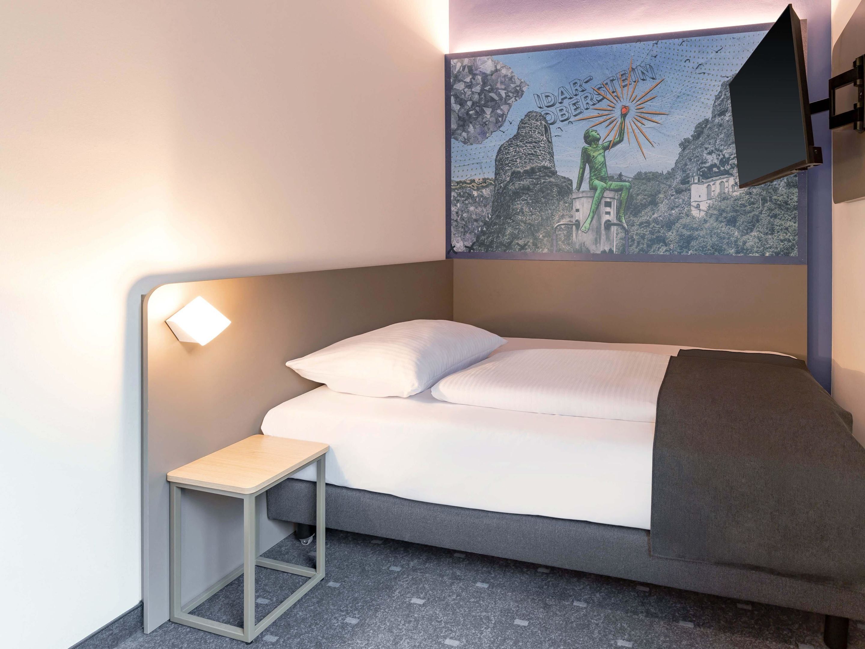 A modern hotel room with a bed, a small bedside table, a wall-mounted TV, and a painting of a landscape.