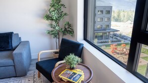 A modern living room with a grey sofa, a dark blue armchair, a small wooden table with magazines, and a large window offering a view of a modern building and playground. - Modern 2 Bedroom Condo with Workspace (Revelstoke)