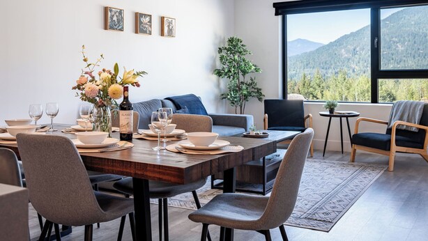 A modern dining area with a wooden table set for four, surrounded by grey upholstered chairs. A large window offers a view of a mountainous landscape. - Modern 2 Bedroom Condo with Workspace (Revelstoke)
