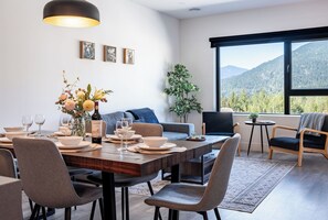 A modern dining area with a wooden table set for four, surrounded by grey upholstered chairs. A large window offers a view of a mountainous landscape. - Modern 2 Bedroom Condo with Workspace (Revelstoke)