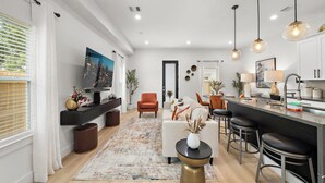A modern living room with a sofa, coffee table, and bar stools. There is a television, a dining area with chairs, and a kitchen area with a counter and cabinets.