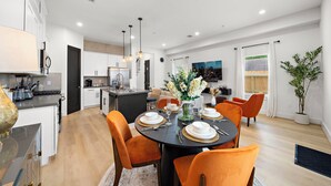 A modern kitchen with white cabinets, a dining area with orange chairs, and a cityscape painting on the wall.