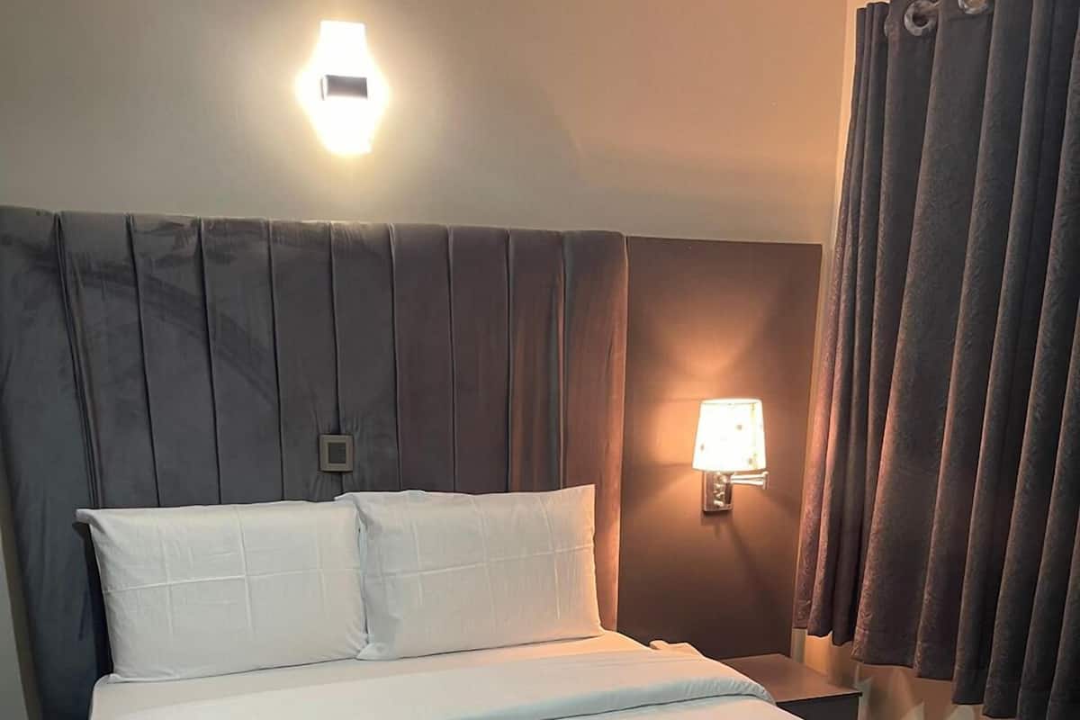 Una cama bien tendida con sábanas blancas, cabecera con paneles verticales, lámpara de noche y cortinas oscuras.