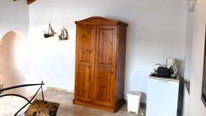 A room with a wooden wardrobe, a small white cabinet with a coffee maker, and a chair with a floral cushion.