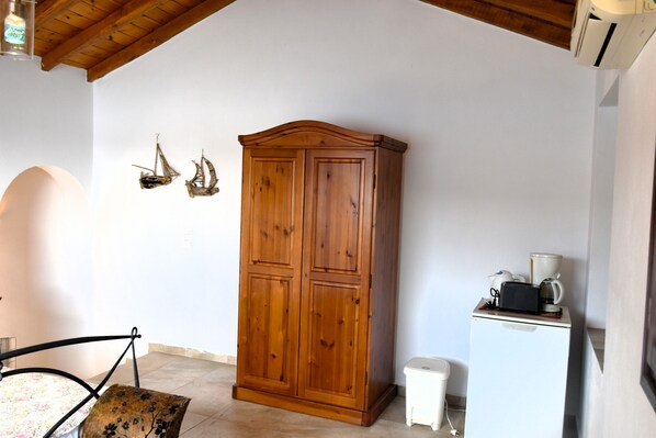 A room with a wooden wardrobe, a small white cabinet with a coffee maker, and a chair with a floral cushion. - Porto Castello Villas Porto Fino Studio (Skiathos)