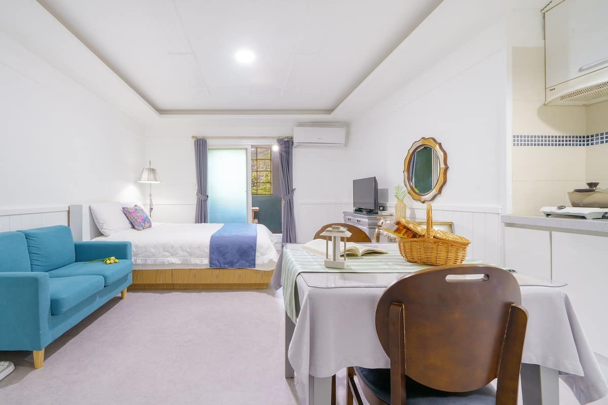 A compact hotel room with a bed, a blue sofa, a dining table with a chair, a television, and a mirror.