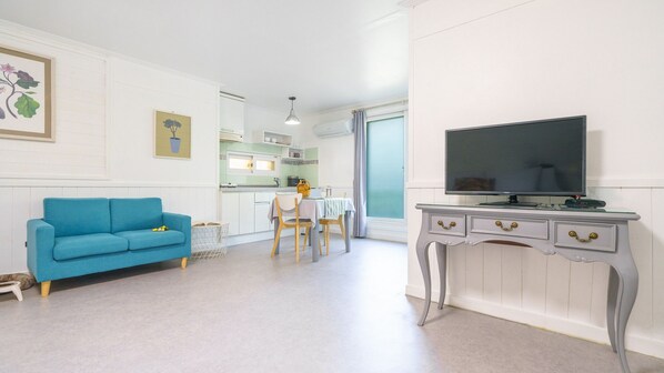 Una sala moderna con un sofá azul, un comedor con mesa y sillas, una cocina con armarios blancos y un televisor sobre una consola blanca.