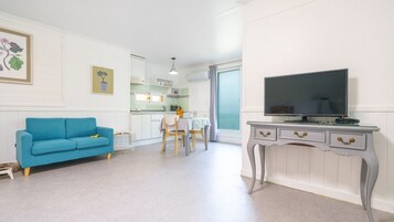 Una sala moderna con un sofá azul, un comedor con mesa y sillas, una cocina con armarios blancos y un televisor sobre una consola blanca.